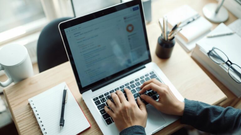 A tidy office desk from above, a laptop with a blurred, unreadable search-results-style interface on the screen, a notebook and a pen beside it, soft morning light through a window – Symbolbild, KI-erzeugt
