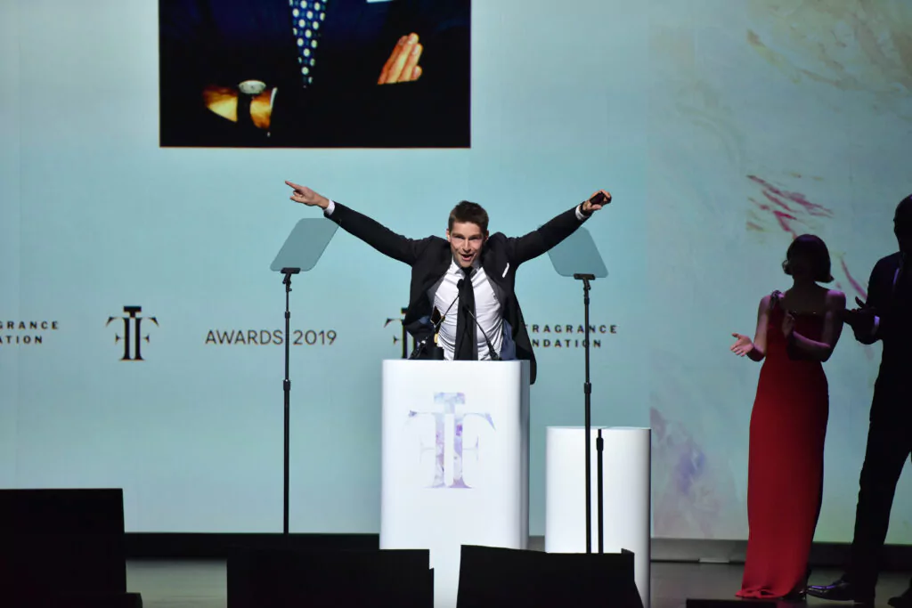 Jeremy Fragrance steht auf einer Bühne und reißt beide Hände in die Höhe. Neben ihm eine Dame mit rotem Kleid. Dahinter ein paar Unternehmenslogos und der Award 2019